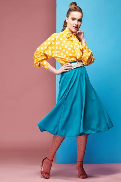 Pretty young sexy serious woman model wearing yellow blouse with white polka-dot, blue skirt and pink tights in pin-up style, posing in studio with blue and pink background — Stock Photo, Image