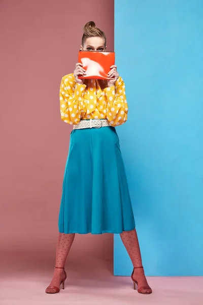 Bastante joven sexy modelo sonriente mujer con blusa amarilla con lunares blancos, falda azul y medias rosadas en estilo pin-up, sosteniendo pequeña bolsa roja y posando en el estudio con fondo azul y rosa —  Fotos de Stock