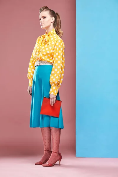 Bastante joven sexy modelo de mujer seria con blusa amarilla con lunares blancos, falda azul y medias rosadas en estilo pin-up, sosteniendo un pequeño bolso rojo y posando en el estudio con fondo azul y rosa —  Fotos de Stock