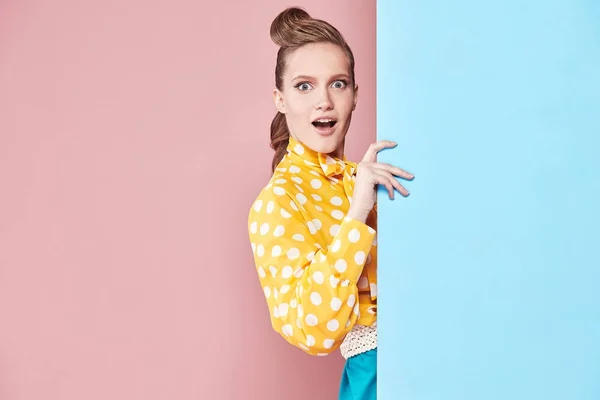 Retrato de atractiva modelo de mujer joven con blusa amarilla con lunares blancos, falda azul en estilo pin-up, posando y mirando sorprendida en estudio con fondo azul y rosa —  Fotos de Stock