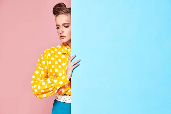 Πορτρέτο του ελκυστική νεαρή γυναίκα σοβαρή μοντέλο φοράει κίτρινο μπλούζα με λευκό πουά, μπλε φούστα σε στυλ pin-up, κρύβονται πίσω από το φως μπλε τοίχο στο studio με μπλε και ροζ φόντο — Φωτογραφία Αρχείου