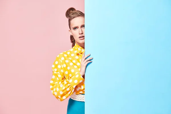 Портрет привабливої молодої серйозної жінки-моделі в жовтій блузці з білими поляками, синя спідниця в стилі пін-ап, ховається за світло-блакитною стіною в студії з синьо-рожевим фоном — стокове фото