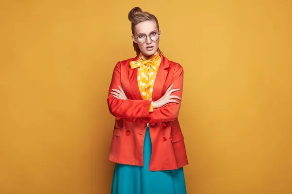 Increíble modelo de mujer seria joven con gafas, con blusa amarilla con lunares blancos, falda azul en estilo pin-up y chaqueta roja, de pie en el estudio sobre fondo amarillo brillante —  Fotos de Stock