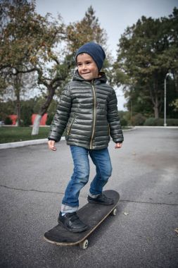 Açık havada gri skate board sürme koyu gri ceket, koyu mavi kap ve mavi jeans, şirin komik küçük boy gülümseyerek  