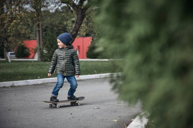 Gri paten kurulu açık havada yeşil ağaçlar arasında sürme koyu gri ceket, koyu mavi kap ve mavi jeans, şirin komik küçük boy gülümseyerek