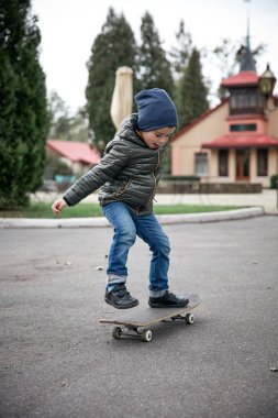 Gri paten kurulu açık havada bir asfalt yolda sürme koyu gri ceket, koyu mavi kap ve mavi jeans, şirin komik küçük boy gülümseyerek
