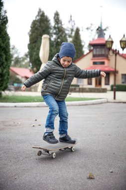 Gri paten kurulu açık havada bir asfalt yolda sürme koyu gri ceket, koyu mavi kap ve mavi jeans, şirin komik küçük boy gülümseyerek