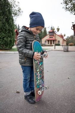 Gri paten holding yönetim kurulu bir asfalt yol üzerinde açık havada koyu gri ceket, koyu mavi kap ve mavi jeans, şirin komik küçük boy gülümseyerek