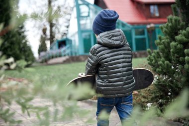 Gri paten kurulu açık havada bir asfalt yol boyunca taşıyan koyu gri ceket, koyu mavi kap ve mavi jeans, şirin komik küçük çocuk