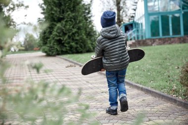 Gri paten kurulu Park'ta bir asfalt yol boyunca taşıyan koyu gri ceket, koyu mavi kap ve mavi jeans, şirin komik küçük çocuk