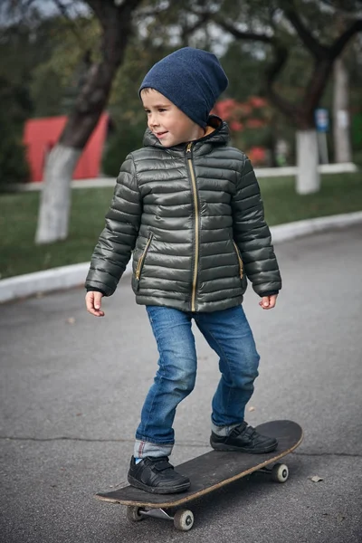 Açık havada gri skate board sürme koyu gri ceket, koyu mavi kap ve mavi jeans, şirin komik küçük çocuk  