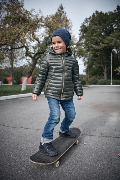 Açık havada gri skate board sürme koyu gri ceket, koyu mavi kap ve mavi jeans, şirin komik küçük boy gülümseyerek  