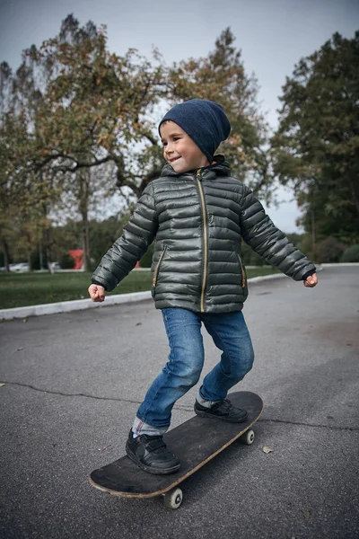 Açık havada gri skate board sürme koyu gri ceket, koyu mavi kap ve mavi jeans, şirin komik küçük boy gülümseyerek  