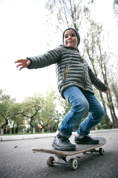 Cute funny smiling little boy wearing dark grey jacket, dark blue cap and blue jeans, riding on grey skate board in garden — Stock Photo, Image