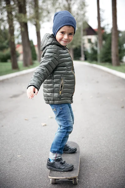 Gri paten kurulu açık havada bir asfalt yolda sürme koyu gri ceket, koyu mavi kap ve mavi jeans, şirin komik küçük boy gülümseyerek