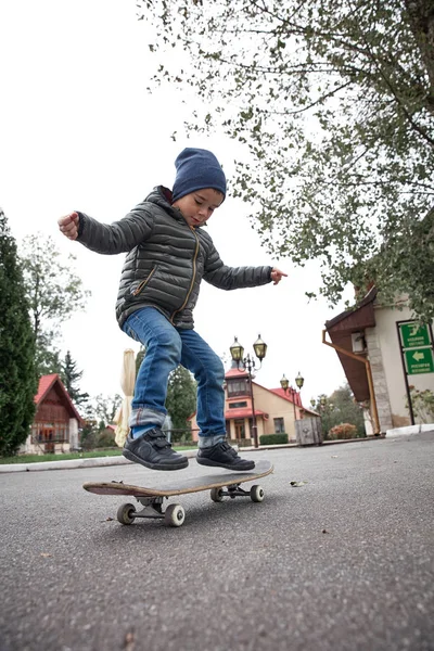 Gri paten kurulu açık havada bir asfalt yolda sürme koyu gri ceket, koyu mavi kap ve mavi jeans, şirin komik küçük boy gülümseyerek