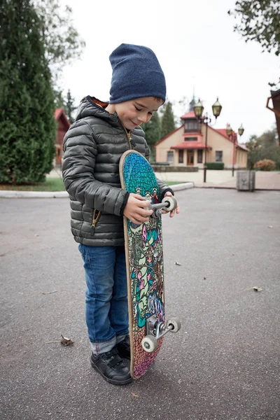 Gri paten holding yönetim kurulu bir asfalt yol üzerinde açık havada koyu gri ceket, koyu mavi kap ve mavi jeans, şirin komik küçük boy gülümseyerek