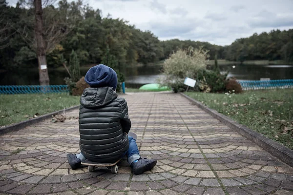 Schattige kleine dappere jongen dragen van donkere grijze jas, donker blauw cap en blue jeans, rijden op grijs skate board in park — Stockfoto