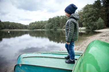 Koyu renk ceketli, mavi kot pantolon, siyah ayakkabı ve göl kıyısında gemide duran koyu mavi şapka giyen küçük şirin çocuk 