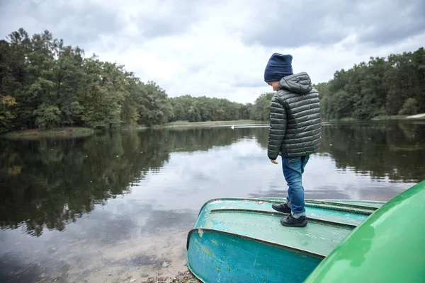 Anak kecil yang lucu mengenakan jaket hitam, celana jeans biru, sepatu hitam dan topi biru gelap, berpose di perahu di tepi danau — Stok Foto