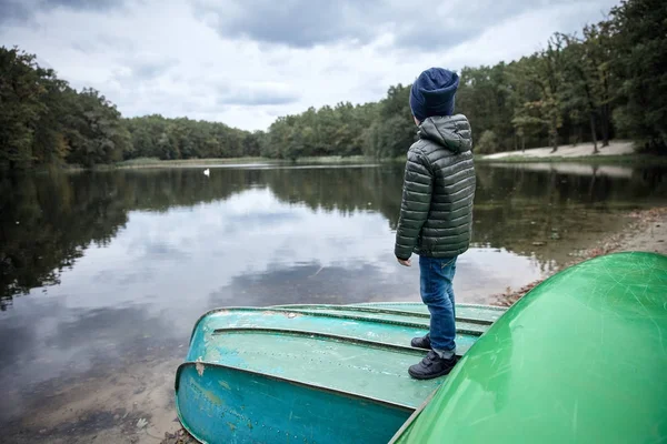Anak kecil yang lucu mengenakan jaket gelap, jeans biru, sepatu hitam dan topi biru tua, berdiri di atas perahu di tepi danau — Stok Foto