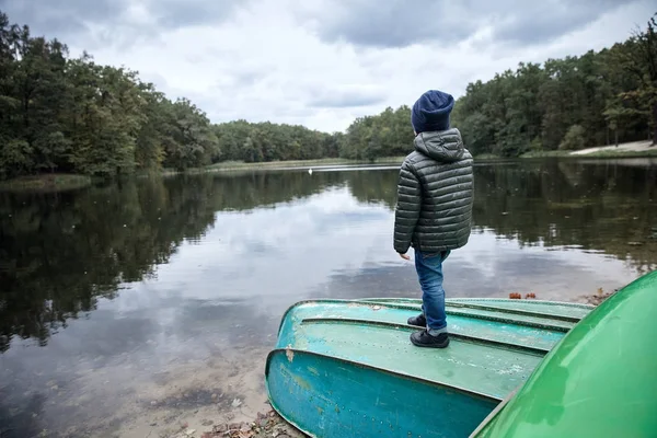 Anak kecil yang lucu mengenakan jaket gelap, jeans biru, sepatu hitam dan topi biru tua, berdiri di atas perahu di tepi danau — Stok Foto