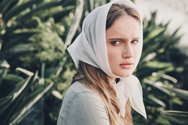 Portrait of young beautiful serious girl with long brown hair wearing light gray dress in retro style and white scarf on her head, posing in greenhouse full of plants — Stock Photo, Image