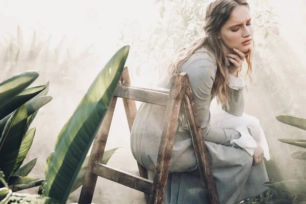 Jovem menina muito séria com cabelos castanhos compridos vestindo vestido cinza claro em estilo retro, posando em escada de madeira em estufa cheia de plantas e sol Imagens De Bancos De Imagens