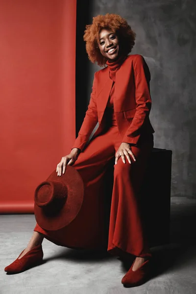 Amazing young black smiling woman with red hair wearing red wide pants, jacket, sweater and shoes sitting in studio with grey and red background and holding red hat