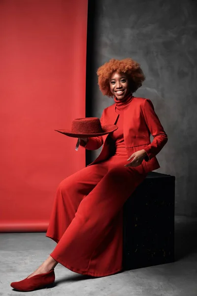 Jolie jeune femme souriante noire aux cheveux roux portant un pantalon large rouge, une veste, un pull et des chaussures assises en studio avec un fond gris et rouge avec un chapeau rouge Photos De Stock Libres De Droits