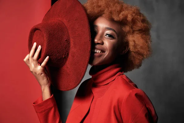 Portrait of beautiful sexy smiling black female model wearing red sweater and jacket, holding red hat in her hand in studio with grey background