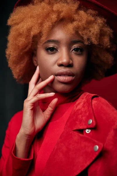 Potret model wanita kulit hitam yang cukup seksi mengenakan sweater merah dan jaket, dengan rambut keriting merah dan topi merah di studio dengan latar belakang abu-abu — Stok Foto