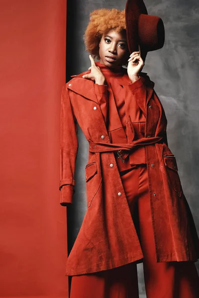 Wanita muda hitam seksi cantik dengan rambut pendek keriting merah mengenakan celana merah, jaket dan sepatu, memegang topi di tangan, berpose di studio dengan latar belakang merah dan abu-abu — Stok Foto