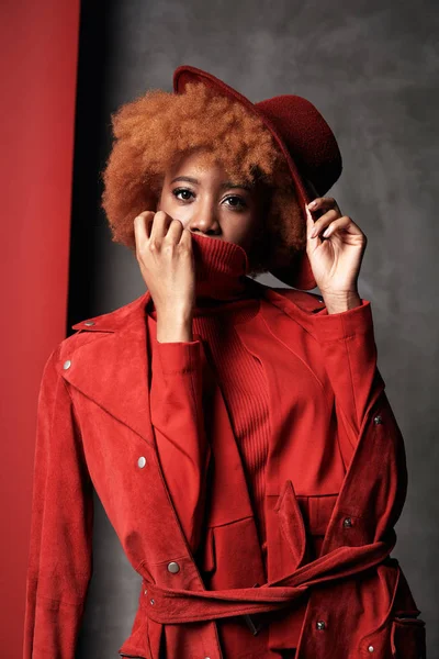 Wanita muda kulit hitam cantik seksi dengan rambut pendek keriting merah mengenakan celana merah dan jaket, memegang topi di tangan, berpose di studio dengan latar belakang merah dan abu-abu — Stok Foto