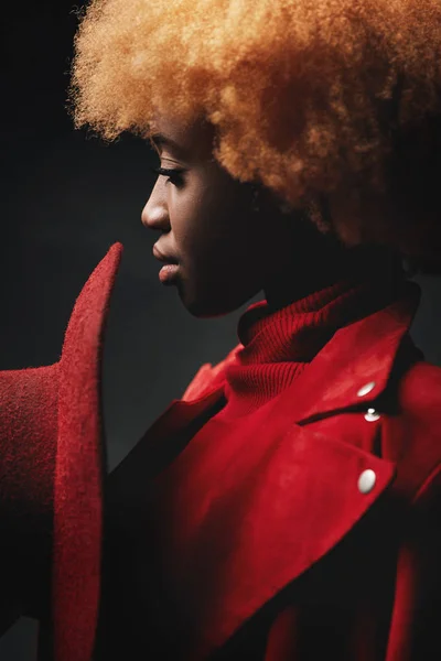 Potret wanita muda dengan model serius seksi dengan rambut pendek keriting merah mengenakan jaket merah, memegang topi merah dan berpose di studio dengan latar belakang gelap — Stok Foto