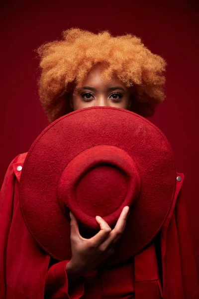 Portrait of young pretty black sexy serious model woman with red curly short hair wearing red sweater, holding red hat and posing in studio on red background