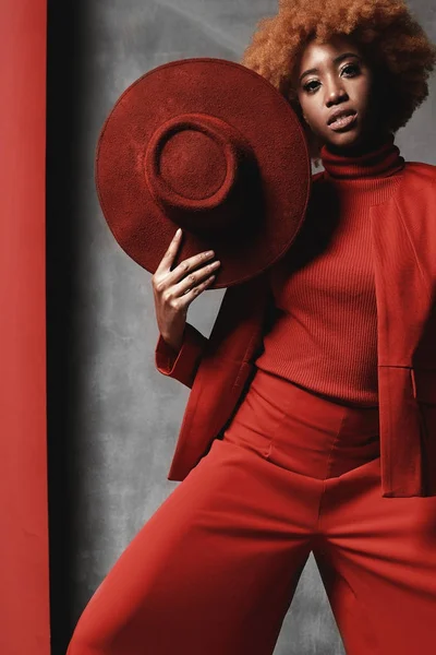 Muda cukup hitam seksi wanita serius mengenakan celana merah, jaket dan sepatu, memegang topi di tangan, berpose di studio dengan latar belakang merah dan abu-abu — Stok Foto