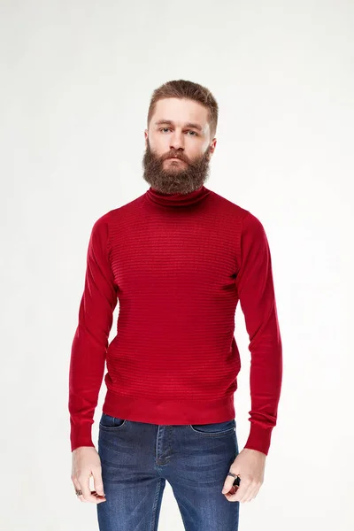 Привлекательный молодой сексуальный серьезный мужчина-модель с бородой и темными волосами позирует в теплом красном свитере и темно-синих брюках в светлой студии — стоковое фото