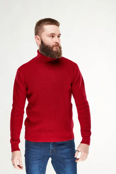 Привлекательный молодой серьезный мужчина-модель с бородой и темными волосами позирует в красном свитере и темно-синих брюках в светлой студии — стоковое фото