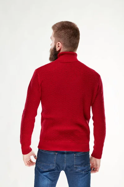 Привлекательный молодой серьезный мужчина модель с темными волосами и бородой позирует в красном свитере и темно-синих брюках в светлой студии — стоковое фото