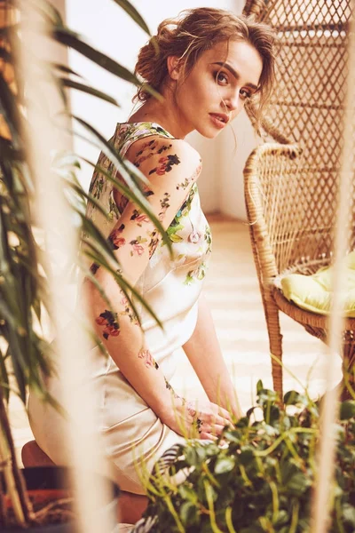 Potret cerah wanita seksi muda yang menakjubkan dengan gaya rambut updo dan tato bunga di lengan dan bahunya mengenakan gaun bunga sutra di studio dengan tanaman dan kursi jerami — Stok Foto