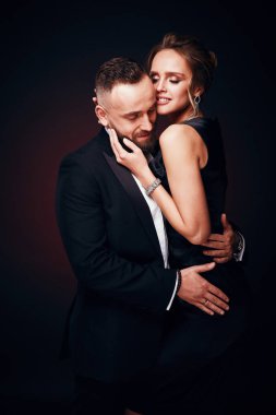 Seksi, lüks çift: yakışıklı adam smokin ile ipeksi siyah elbise ve şık takı, karanlık Studio'da poz giyen inanılmaz kadın sarışın kabarık saçlı sakallı