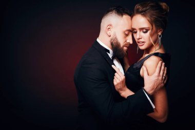 Lüks, şık çift: yakışıklı adam smokin ile ipeksi siyah elbise ve şık takı, karanlık Studio'da poz giyen inanılmaz kadın sarı saçlı sakallı