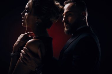 İpeksi siyah elbise ve şık takı ve yakışıklı sakallı adam ile harika bir smokin giyiyor tutkulu kadın sarışın kabarık saçlı koyu Studio'da poz