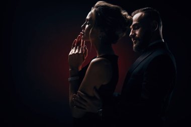 Şık çift: sarışın kabarık saçlı kadın ipeksi siyah elbise ve lüks mücevher ve yakışıklı sakallı adam ile harika bir smokin karanlık Studio'da poz giyiyor