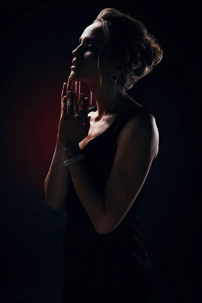 Dark portrait of amazing blonde sexy woman with glamorous makeup, wearing silky black dress and chic jewelry, posing in studio with dark background