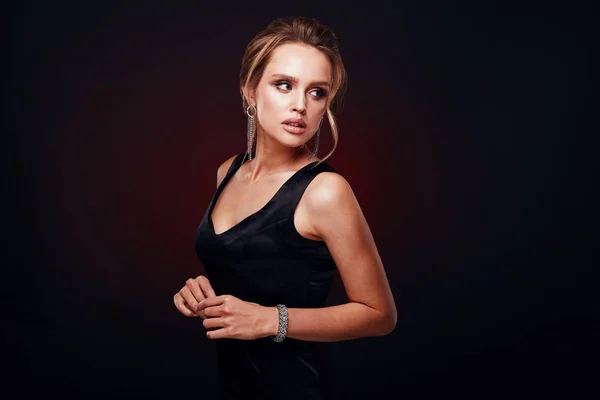 Amazing sexy woman with blonde updo hair and glamorous makeup, wearing silky black dress and chic jewelry, posing in studio with dark background — Stock Photo, Image