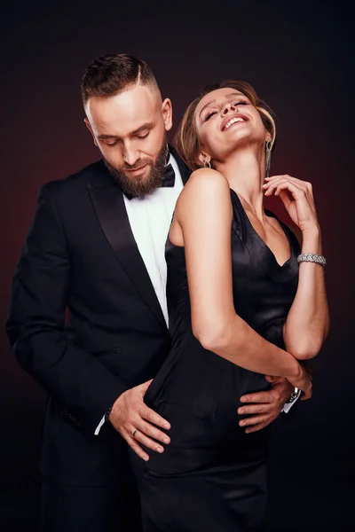 Casal luxuoso, elegante e feliz: homem barbudo bonito em smoking com mulher incrível com cabelo loiro updo, vestindo vestido preto sedoso e jóias chiques, posando em estúdio escuro — Fotografia de Stock