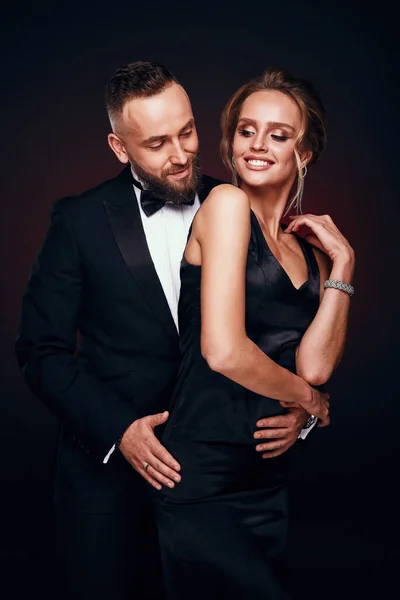 Casal luxuoso e elegante: homem barbudo bonito em smoking com mulher incrível com cabelo loiro updo, vestindo vestido preto sedoso e jóias chiques, posando em estúdio escuro — Fotografia de Stock