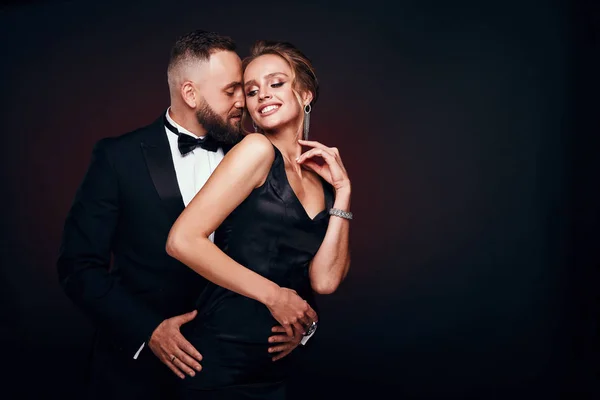 Casal luxuoso e elegante: homem barbudo bonito em smoking com mulher sorridente incrível com cabelo loiro updo, vestindo vestido preto sedoso e jóias chiques, posando em estúdio escuro — Fotografia de Stock